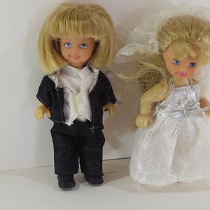 May include: A pair of small wedding dolls, a bride and groom, with blonde hair and blue eyes. The bride is wearing a white lace dress with a veil, and the groom is wearing a black suit with a white shirt and vest.