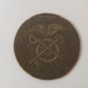 May include: A circular, olive-drab patch featuring an embroidered design. The design includes a spread-winged eagle above crossed keys and a wheel. The patch is likely a military insignia or emblem, possibly for a specific unit or role.