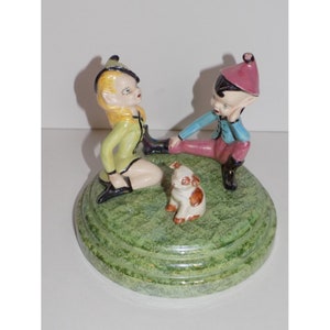 May include: A ceramic figurine set featuring two elves sitting on a green base with a small white and brown dog between them. The female elf is wearing a green and yellow outfit with a black hat, while the male elf is wearing a blue and pink outfit with a red hat.