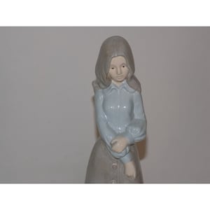 May include: A porcelain figurine of a woman wearing a light blue dress and a gray hooded cloak. She has long brown hair and is holding a small child in her arms.