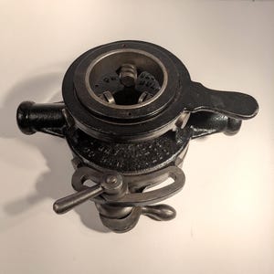 May include: A black cast iron pipe threading tool featuring a circular die head with internal metal teeth, a side-mounted handle, and an adjustable locking lever mechanism. The body displays embossed text including the words OSTER and BULL DOG.