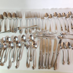 May include: A set of silverware, including spoons, forks, and knives, all made of polished stainless steel. The silverware is arranged in rows on a white surface.