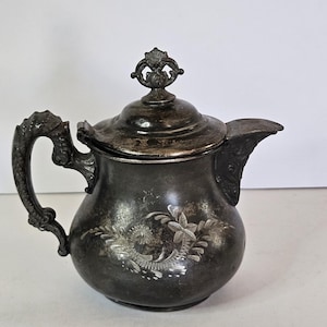 May include: Antique silver-toned teapot with a decorative floral design. The teapot features a curved handle, a spout, and a lid with a decorative finial. The body of the teapot has an embossed floral pattern.