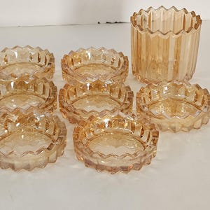 May include: A set of seven amber-colored glass ashtrays with a decorative, scalloped edge, and a matching cylindrical vase. The ashtrays are round and have a starburst pattern in the center. The vase has vertical ridges.