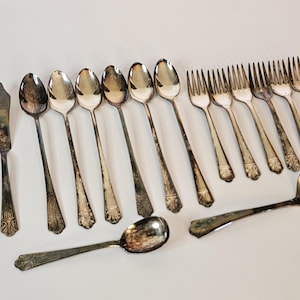 May include: An assortment of antique silverware, including spoons, forks, and a butter knife, arranged on a white surface. The silverware has a tarnished, vintage appearance, with decorative detailing on the handles. A soup ladle is also included.