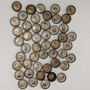 May include: A collection of numerous metal bottle caps, each featuring a small circular black and white portrait of a different individual. The caps have serrated edges and show varying degrees of age, patina, and metallic wear across the set.
