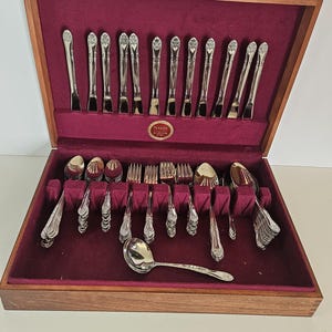 May include: A wooden storage chest with a deep burgundy velvet interior containing a set of silver-toned flatware. The set includes knives, forks, and spoons with ornate handles. A small oval label inside reads Naken Silverware Chest.