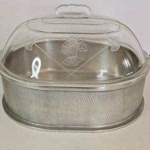 May include: An oval-shaped metal casserole dish with a textured exterior and two side handles. It includes a clear glass lid featuring an etched design of a knight's helmet centered between two crossed halberds, topped with a glass handle.