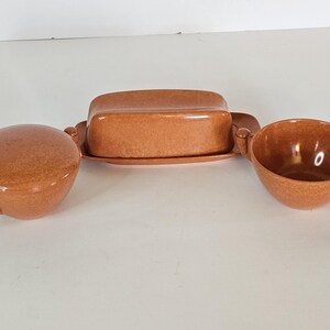 May include: A three-piece ceramic serving set in a speckled brown finish. The set includes a covered butter dish, a creamer, and a sugar bowl with a lid. The pieces have a mid-century modern design.
