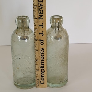 May include: Two vintage clear glass bottles with rounded shoulders and narrow necks. A wooden ruler with black markings and the text "Compliments of J. J. Newe" is placed next to the bottles. The bottles are approximately 6 inches tall.