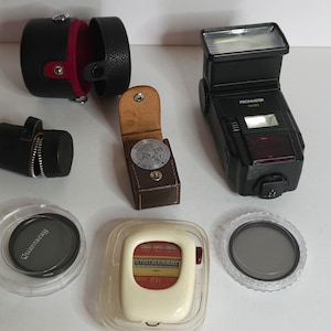 May include: A collection of photography accessories including a black flash unit labeled "Promaster 1400", a black leather case with a zipper, a brown leather case with a light meter, a white light meter, a black filter, and a clear filter.