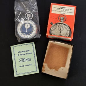 May include: A silver stopwatch with a blue face inside a clear plastic bag, an orange box labeled Compass Instrument & Optical Co., Inc. Swiss Stop Watch, and a light green Certificate of Guarantee card with a brown-lined empty box base.