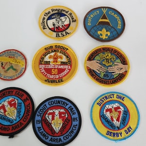 May include: A collection of eight embroidered scout patches. The patches are circular and feature various designs, including a scout handshake, a tent, a diamond, and a scout emblem. The patches are in various colors, including blue, yellow, red, and green.