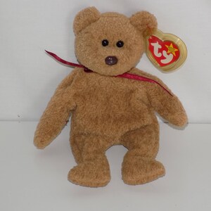 May include: A brown Ty Beanie Baby bear with a red ribbon around its neck. The bear has a brown nose and black eyes. The bear is wearing a Ty Beanie Baby tag with a red heart and the word "Ty" on it.
