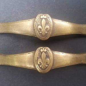May include: Two antique brass drawer pulls with a fleur-de-lis design. The pulls are oval shaped with a raised design and have a smooth, polished finish.