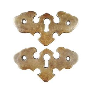 Könnte beinhalten: Zwei antike Schlüsselschilder aus Messing mit filigranen Designs. Jedes Schild hat ein Schlüsselloch in der Mitte und zwei dekorative Löcher an den Seiten.