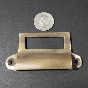 May include: Brass drawer pull with a rectangular label holder. The pull has a rounded edge and two small holes on either side.