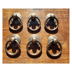 May include: Six antique brass ring cabinet pulls with a rounded top and a circular ring handle. The pulls are arranged in two rows of three on a wooden surface.