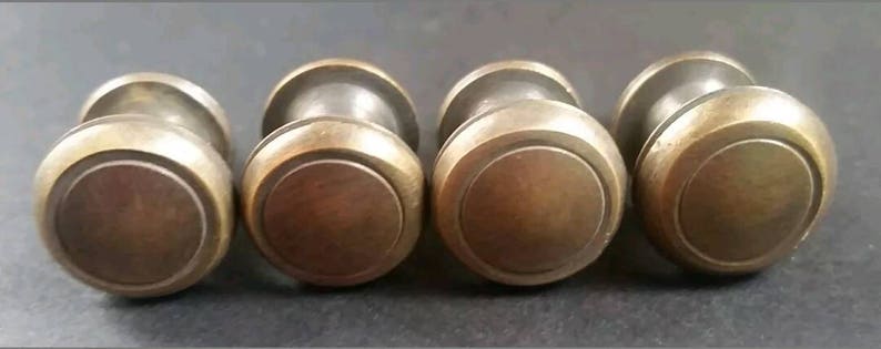 May include: Four antique brass cabinet knobs with a round, slightly raised, centre design. The knobs have a smooth, polished finish.