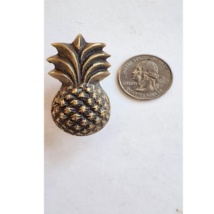 Op de afbeelding: Een gouden ananasvormige kastknop met een gedetailleerd ontwerp. De knop is ongeveer zo groot als een Amerikaanse kwart.