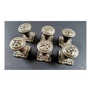 May include: Six antique brass cabinet knobs with a square base and a round top with an ornate design.