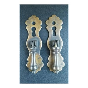 Peut inclure: Deux poignées de tiroir en laiton antique avec un motif de trou de serrure et un bord festonné décoratif et orné. Les poignées ont une finition foncée vieillie et un design simple et élégant.