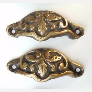 May include: Two antique brass drawer pulls with a floral design. The pulls are shaped like a half-circle and have two holes for mounting.