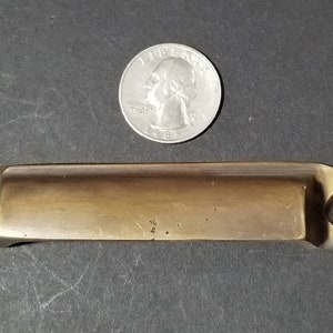 May include: A brass door pull with rounded edges and two holes for mounting. The pull is about 2 inches long and 1 inch wide.