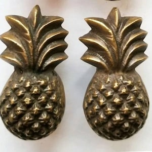 May include: Two gold pineapple-shaped drawer pulls. The pineapple is detailed with a textured surface and a leafy top.