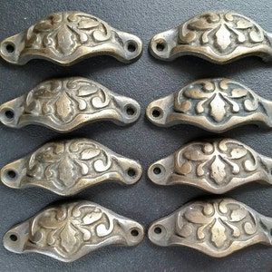 May include: Eight antique brass drawer pulls with ornate floral designs. Each pull has a curved shape and two small holes for mounting.
