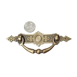 Pode incluir: Uma puxador de gaveta de latão com um design ornamentado. O puxador tem um círculo central com um design em relevo e uma alça curva.