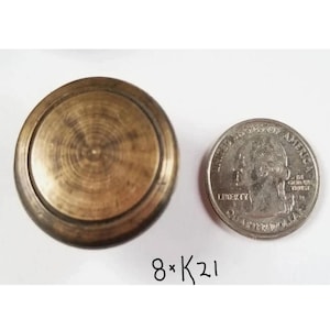 May include: A round, brass knob with a raised edge and a smooth, shiny surface. The knob is about the size of a US quarter.