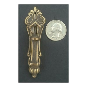 May include: A brass cabinet pull with an ornate design. The pull is about the size of a 25 pence coin.