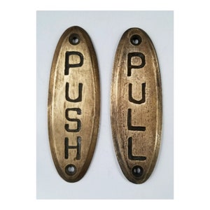 May include: Two oval-shaped brass door signs with black lettering. The sign on the left reads "PUSH" and the sign on the right reads "PULL".