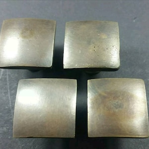 May include: Four square-shaped brass cabinet knobs with a slightly rounded edge. The knobs have a brushed finish and are a light gold colour.