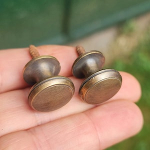 May include: Two antique brass cabinet knobs with a round, slightly raised, decorative edge. The knobs have a screw on the back for attaching to a cabinet door.