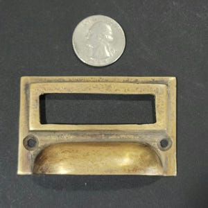 May include: A brass drawer pull with a rectangular label holder. The pull has a curved, recessed handle and two small, round holes on either side.