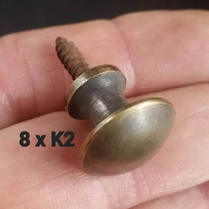May include: A close-up of a brass knob with a screw. The knob is round and has a slightly domed top. The screw is threaded and has a slightly rounded head. The knob is a light brass colour and has a slightly aged look. The text "8 x K2" is visible in the image.