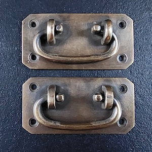 Könnte beinhalten: Zwei antike Messing-Schranktürgriffe in rechteckiger Form mit einem abgerundeten, gewölbten Griff. Die Griffe haben eine abgenutzte, gealterte Oberfläche.