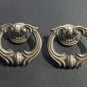 Puede incluir: Dos tiradores de armario de latón adornados con un asa de anillo y volutas decorativas. Los tiradores son de estilo tradicional y tienen un aspecto vintage.