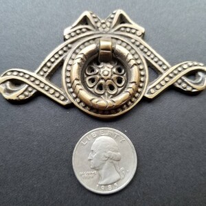 May include: A brass cabinet pull with a decorative, ornate design. The pull features a ring handle and a circular base with a floral pattern. The pull is approximately the size of a US quarter.