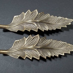 Peut inclure: Deux pièces décoratives en forme de feuille en laiton orné. Les feuilles sont détaillées avec des lignes en relief et ont une surface légèrement texturée.