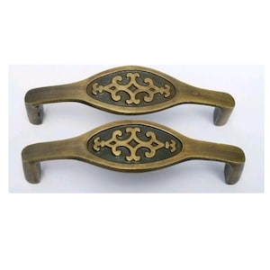 Puede incluir: Dos tiradores de armario de latón antiguo con un diseño ornamentado. Los tiradores son de forma ovalada con una sección central elevada que presenta un patrón geométrico.