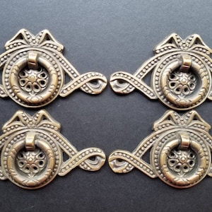 May include: Set of four antique brass drawer pulls with ornate designs, featuring a ring pull and a decorative bow at the top.