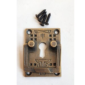 May include: Antique brass cabinet door latch with a keyhole and decorative details. The latch has two small knobs on either side of the keyhole. The latch is accompanied by four black screws.