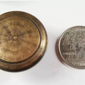 May include: A round, brass knob with a raised edge and a concentric circle pattern. The knob is about the size of a US quarter.