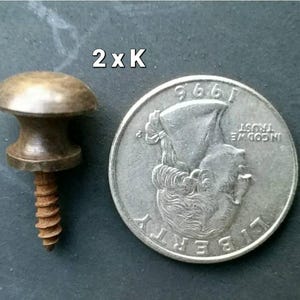 Puede incluir: Primer plano de un pomo de latón con un tornillo. El pomo es del tamaño aproximado de una moneda de 25 centavos de dólar estadounidense. El texto "2 x K" es visible en la imagen.