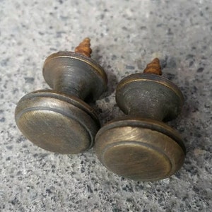 Op de afbeelding: Twee antieke messing kastknoppen met een afgeronde bovenkant en een enkele schroef. De knoppen hebben een versleten, verweerde afwerking.