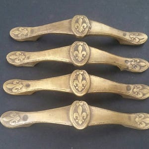 May include: Four antique brass drawer pulls with a fleur-de-lis design. Each pull has a decorative, ornate design with a raised fleur-de-lis in the center.