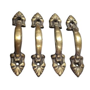 May include: Four antique brass cabinet door pulls with ornate designs. Each pull has a curved handle and a decorative, pointed end.